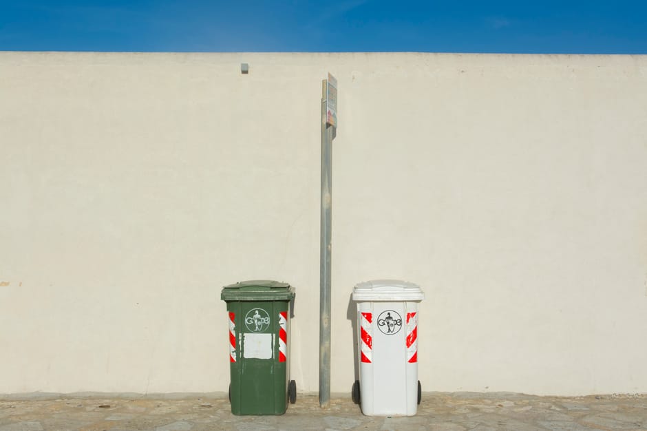 Two wheelie bins, one green and one white, are positioned side by side against a plain cream-colored wall under bright daylight. The green bin, on the left, features a recycling symbol and is made of 