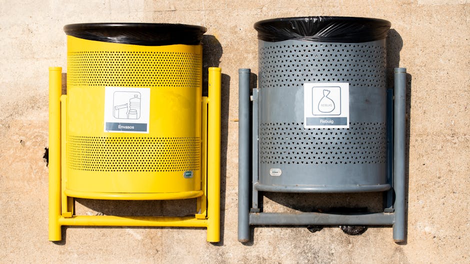 Two wheelie bins, one green and one white, are positioned side by side against a plain cream-colored wall under bright daylight. The green bin, on the left, features a recycling symbol and is made of 