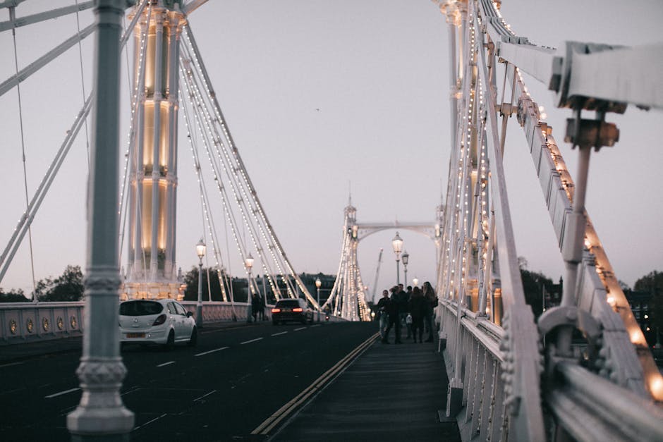 The image depicts the Hammersmith Bridge during dusk, with its white suspension cables and structure illuminated by warm string lights. The bridge's walkway, lined with classic lamp posts, appears cle