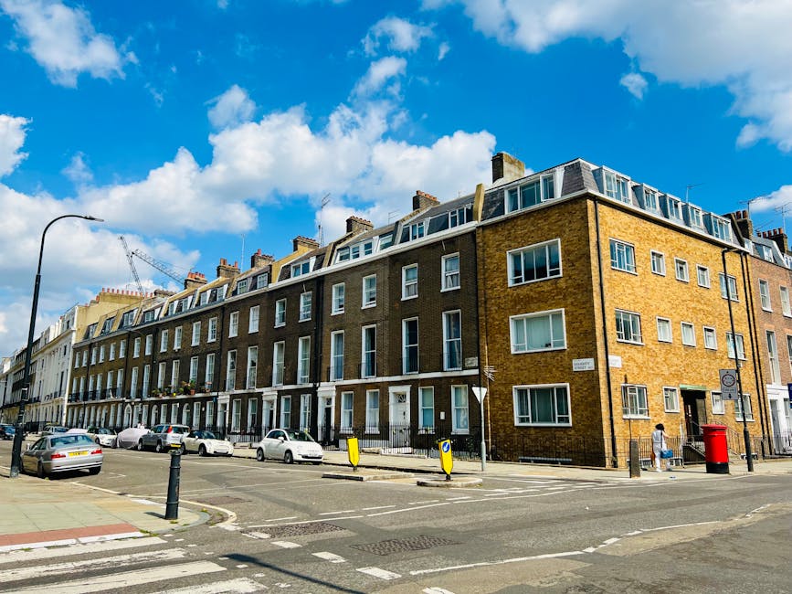 A row of multi-story residential buildings with brown brick facades, white window frames, and pitched roofs under a bright blue sky with scattered clouds in Hammersmith. The buildings are situated alo